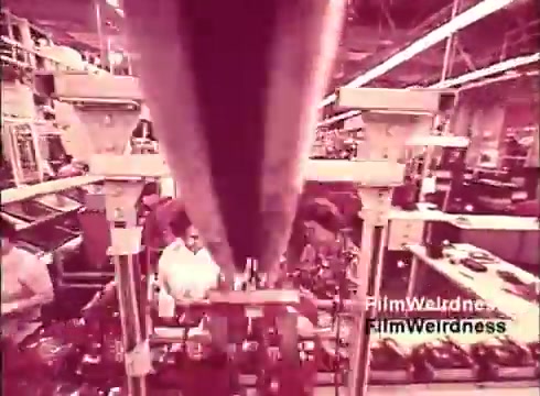 The still from 'Bell Performance' captures a manufacturing scene within a factory, likely from the 1970s, showing the production of telephones. The image focuses on assembly equipment and workers, emphasizing a nostalgic glimpse into the industrial process of creating communication devices. The visual tone suggests a vintage aesthetic, enhancing the sense of history associated with the Bell System.