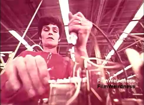 The still from the 1970s film 'Bell Performance' showcases a technician working on telephone equipment in a manufacturing environment. The focus is on a woman using tools to assemble or repair a device, highlighting the hands-on nature of telephone production. The color palette is likely muted or vintage, reflective of the era, adding to the nostalgic feel of Bell System's history in telecommunications. The absence of titles suggests a functional, documentary style aimed at illustrating the intricacies of telephone manufacturing.