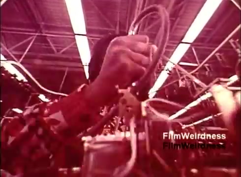The still from the 'Bell System Film from the 1970s' showcases a close-up of a worker's hand engaging with telephone wiring in a factory setting. The atmosphere is nostalgic, emphasizing the craftsmanship involved in telephone production. The warm, reddish tint of the film adds to the vintage feel, evoking a sense of history associated with the Bell System. This moment captures the intricate and manual nature of electronics assembly during that era.