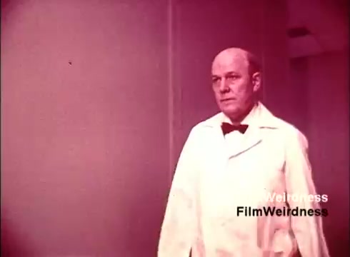 The still depicts a man in a white lab coat and a bow tie, walking through a sterile environment, likely a production or laboratory setting. The overall color tone has a warm, reddish hue, giving it a nostalgic feel typical of 1970s films. This scene suggests a focus on the meticulous processes behind telephone manufacturing, evoking a sense of professionalism and innovation associated with the Bell System.