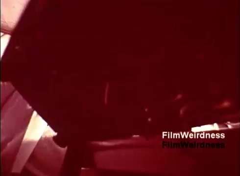 The still from the film 'Bell Performance' showcases a close-up view of machinery, possibly related to the production of telephones. The image has a reddish tint, which adds a vintage feel typical of 16mm films from the 1970s. The focus appears to be on the inner workings of the manufacturing process, highlighting the intricate components involved in creating telephones. The absence of titles suggests a straightforward, documentary-style approach to capturing the nostalgia of Bell System's operations.