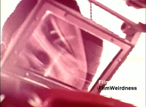 The still from the 16mm film 'Bell Performance' captures a close-up view of a person interacting with a telephone component, possibly during its assembly or inspection. It features a reflective surface, creating a distorted visual effect. The colors appear warm, suggesting a nostalgic tone, and the context hints at the mid-20th century technology era, emphasizing the craftsmanship behind telephone manufacturing. The overall aesthetic evokes a sense of curiosity about the Bell System's history and innovations.