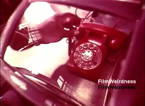 The still from the film 'Bell Performance' shows a close-up of a red rotary telephone, reflecting a metallic surface. The image captures a nostalgic moment, highlighting the simplicity and design of mid-20th century telecommunication devices. The background suggests a manufacturing or assembly environment, indicative of the era's focus on craftsmanship in technology. Overall, it's a snapshot that evokes nostalgia for the Bell System and its iconic products from the 1970s.