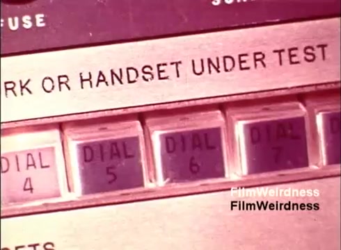 The still from the 16mm film titled 'Bell Performance' showcases an industrial telephone testing apparatus from the 1970s. The focus is on a control panel featuring multiple buttons labeled with 'DIAL' and corresponding numbers. The panel is marked 'MARK OR HANDSET UNDER TEST,' suggesting a quality assurance or testing process for telephones. The aesthetic reflects the technology and design of that era, evoking a sense of nostalgia related to the Bell System and the evolution of telephone technology.