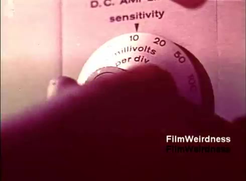 The still features a close-up of a hand adjusting a control knob on a device labeled for measuring sensitivity in millivolts. This scene likely captures a moment focused on technical craftsmanship related to telecommunication, emphasizing the precision involved in telephone manufacturing during the 1970s. The film reflects a nostalgic look at the Bell System, showcasing both the technology and the hands-on work that went into creating communication devices of that era.