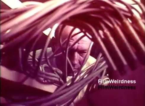 The still from 'Bell Performance' features a close-up of a man surrounded by a tangle of wires, likely in a manufacturing or assembly environment related to telephones. The lighting suggests a vintage aesthetic typical of the 1970s, contributing to the overall nostalgic feel of the film. The man's focused expression conveys his engagement with the intricate work involved in the creation of telecommunications equipment.