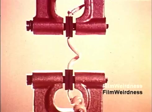 The still from 'Bell System Film' features a close-up of two large, metallic structures that appear to be part of a telephone assembly process. A coiled telephone wire connects the two components, emphasizing the intricate mechanics behind telephone technology. The image captures a nostalgic glimpse into the manufacturing of telephones, highlighting both the engineering and the aesthetic of the era. The muted colors lend a vintage feel to the scene, reinforcing its historical context.