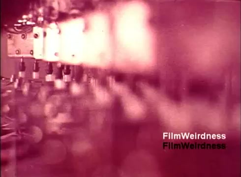 The still captures a close-up view of telephone manufacturing equipment from the 1970s, showcasing intricate machinery and components involved in the production process. The color palette leans towards a reddish hue, giving the image a nostalgic feel. The focus is soft, creating a dreamy quality, while various components and connections hint at the complexity of telephone technology during that era. The text 'FilmWeirdness' is superimposed, adding a contemporary layer to this historical snapshot.