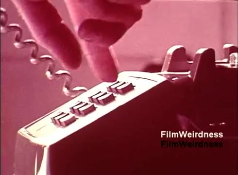 The still captures a close-up of a hand poised to press buttons on a vintage telephone, indicative of 1970s design. The telephone appears to be a push-button model, reflecting the era’s shift from rotary dials. The background hints at a production environment, emphasizing the manufacturing process. The film, part of Bell System nostalgia, highlights the intricacies of telephone technology during that time.