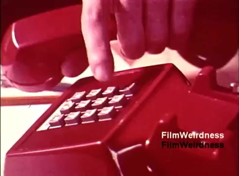 The still from the 1970s film 'Bell Performance' features a close-up of a red rotary telephone. A hand is poised above the phone, ready to dial, highlighting the keypad's classic design. This moment captures the nostalgia of telecommunication evolution, reflecting the era's technology and aesthetic. The film, devoid of titles, immerses viewers in Bell System history, showcasing the process of telephone creation and operation.