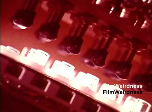 The still from the 16mm film 'Bell Performance' captures an intriguing close-up of a telephone assembly line. It features various components, likely switches or circuit elements, against a backdrop of warm illumination. This visual emphasizes the intricate manufacturing process of telephones, providing a nostalgic glimpse into the technology of the 1970s and the Bell System's role in telecommunications. The overall tone is one of industrial efficiency, showcasing both the complexity and craftsmanship involved in creating these devices.