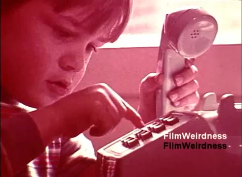 The still from the 16mm film 'Bell Performance' captures a nostalgic moment, featuring a young boy intently interacting with a vintage telephone. His focus on pressing the buttons highlights the era's connection to communication technology. The warm tones evoke a sense of nostalgia, reflecting the influence of the Bell System during that time. Overall, the image conveys a blend of childhood curiosity and the historical significance of telephony.