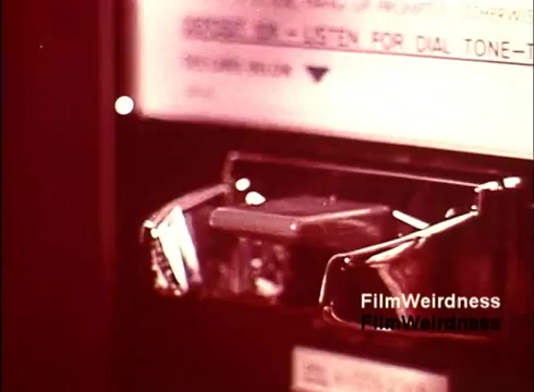 The still from the 16mm film 'Bell Performance' showcases a close-up of a telephone booth, highlighting the intricate mechanisms typically found within. The shot captures the details of the dialing system, focusing on the rotary dial and its associated components. The color scheme suggests a vintage aesthetic, reflective of the 1970s era. Overall, the image evokes a sense of nostalgia for the time when telephones were fundamental communication tools.