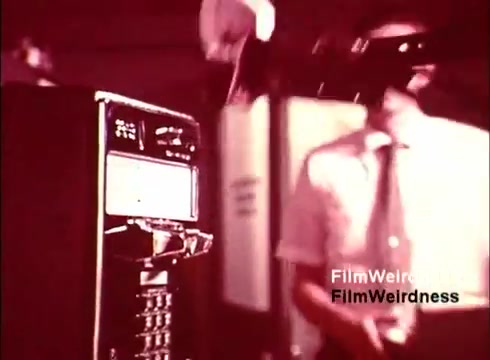 The still from 'Bell Performance' features a vintage scene depicting the production of telephones. In the foreground, a payphone unit is prominently displayed, while a worker in a light-colored shirt and tie is engaged in assembling or manipulating components nearby. The setting appears to be a factory or workshop, highlighting Bell System's technology from the 1970s, evoking a sense of nostalgia for the era and its advancements in telecommunications. The colors are likely warm and muted, typical of film from that time period.