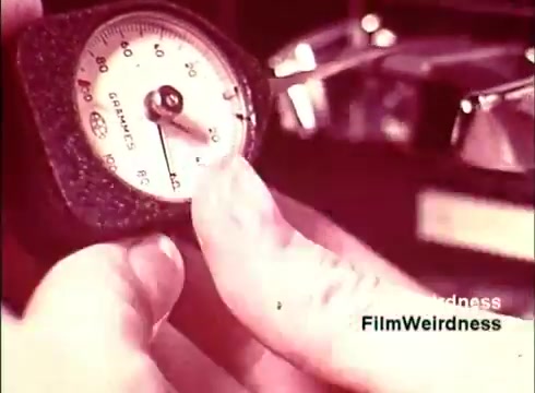 The still from the 'Bell System Film from the 1970s' features a close-up of a hand adjusting a gauge, likely measuring some parameter in the manufacturing process of telephones. The background suggests a workshop environment, possibly depicting quality control or testing procedures. The overall aesthetic evokes a sense of nostalgia for the era of telecommunications development.