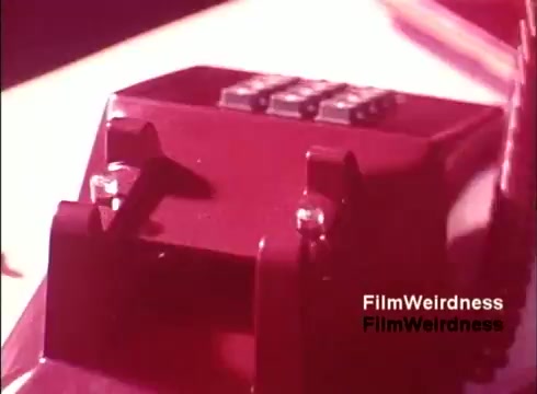 The still shows a close-up of a red telephone from the 1970s, likely emphasizing its design and features. The film, titled 'Bell Performance,' captures the manufacturing process of telephones, showcasing Bell System nostalgia. The focus on the device suggests an exploration of communication technology during that era. The colors used provide a retro aesthetic typical of the time.