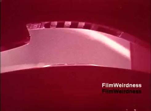 The still from the 'Bell Performance' film showcases a close-up view of film in a projector, likely highlighting the process of filmmaking. The red tint adds a nostalgic effect, imbuing it with a vintage aesthetic typical of the 1970s. The film may capture scenes of telephone manufacturing or technology associated with the Bell System, reflecting a bygone era of telecommunication innovation.