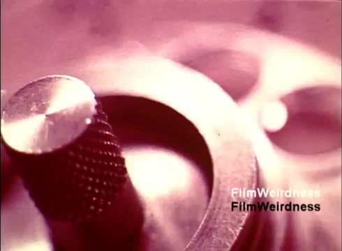 The still from the 'Bell System Film from the 1970s,' titled 'Bell Performance,' features a close-up of industrial machinery, likely related to telephone manufacturing. The image highlights a metal component, showcasing intricate details and textures, which suggests precision engineering. The film captures the nostalgic essence of the Bell System and its role in telecommunications history. The color tones are muted, enhancing the vintage feel of the production.