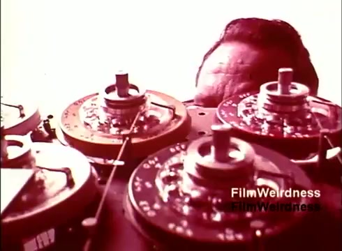 The still from 'Bell Performance' captures a close-up of industrial machinery, likely related to telephone manufacturing. In the foreground, several circular components with dials and knobs are visible, hinting at the intricate technology of the era. A man's head is partially obscured at the top, suggesting he may be operating or inspecting the equipment. The color palette reflects the warm tones typical of 1970s film, enhancing the nostalgic aspect of the scene. Overall, the image conveys a sense of innovation and the behind-the-scenes processes of telecommunication during that time.