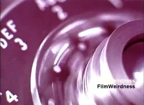 The still features a close-up of a rotary dial from a telephone, showcasing its intricate design and mechanics. The dial is metallic with numbered increments and letters, capturing the vintage aesthetic of telecommunication technology from the 1970s. The overall color tone has a nostalgic quality, evoking memories of an era when such devices were commonplace in households.