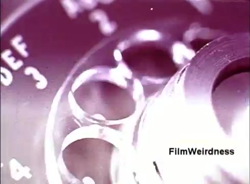 The still captures a close-up of a rotary dial from an old telephone, showcasing its circular design and numbered holes. The image has a nostalgic, vintage aesthetic, likely emphasizing the craftsmanship of telephone manufacturing in the 1970s. The film, titled 'Bell Performance,' reflects on the history and technology of the Bell System, presenting a behind-the-scenes look at telephony. The focus is on the intricate details of the dial, evoking memories of earlier communication methods.