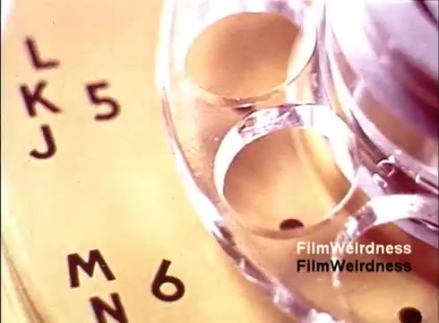 The still from 'Bell Performance' features a close-up of a vintage rotary telephone dial. The golden background highlights the letters 'L,' 'K,' 'J,' 'M,' 'N,' and the numbers '5' and '6' surrounding the dial's finger holes. This nostalgic glimpse reflects the craftsmanship of telephone manufacturing in the 1970s, capturing the essence of Bell System's telecom history. The image evokes a sense of simplicity and innovation from that era.