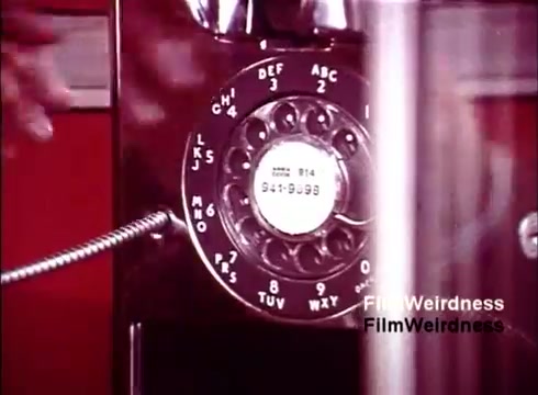 The still features a close-up of a vintage rotary telephone, showcasing its intricate design. The dial includes numbers and letters, indicating its functionality. The background is a striking red, which contrasts with the phone, creating a nostalgic and vibrant atmosphere typical of the 1970s. This image captures a moment in the manufacturing or design process, reflecting the era's telecommunications technology and cultural significance.