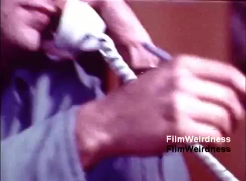 The 16mm still from the film 'Bell Performance' shows a close-up of a person holding a telephone receiver, with a focus on their hands and a portion of the phone. The image captures the craftsmanship involved in creating telephones, reflecting the nostalgic aesthetic of the 1970s era associated with Bell System. The muted colors and composition evoke a sense of industrial history and personal connection.