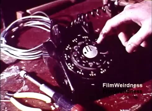 The still captures a close-up of a classic rotary dial telephone, showcasing the intricate details of its design. A hand is seen on the dial, about to turn it, which suggests a focus on the operation of the device. Surrounding the phone are tools and wires, indicating a setting related to telecommunications repair or assembly. The overall composition evokes a sense of nostalgia for the era and the craftsmanship involved in telephone technology during the 1970s.