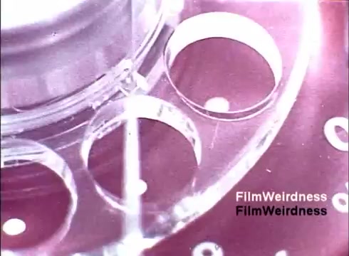 The still captures a close-up view of a rotary dial, showcasing the transparent plastic components and intricate mechanical details. The circular holes for finger placement are prominently featured, highlighting the vintage design typical of telephones from the 1970s. The film has a nostalgic quality, evoking a sense of the era's technology and craftsmanship associated with the Bell System. The muted colors add to its retro aesthetic, emphasizing the charm of outdated communication devices.