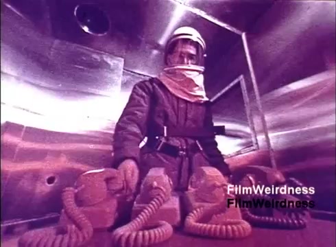 The still from the 1970s 'Bell Performance' film features an individual in a protective suit and helmet, standing in an industrial-like environment. They are surrounded by several vintage telephones with coiled cords, suggesting a focus on the manufacturing process of telecommunications equipment. The scene exudes a nostalgic feel, reflecting the era's fascination with technology and the production of essential communication devices.