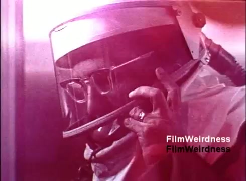 The 16mm still from the 'Bell System Film from the 1970s,' titled 'Bell Performance,' depicts a worker wearing a helmet with a transparent face shield. He is engaged in a call, holding a telephone in one hand. The scene captures the industrial context of telephone manufacturing or technologies associated with the Bell System, evoking nostalgia for the era's telecommunications advancements. The image exudes a vintage aesthetic, characteristic of films from that time.
