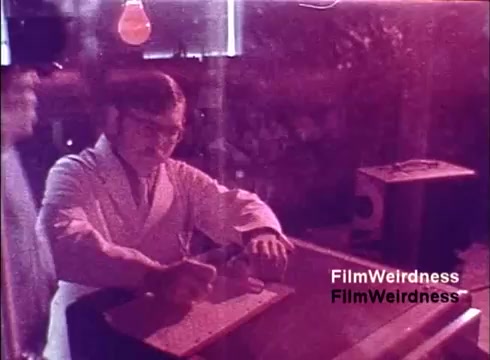 The still from the 1970s Bell System film 'Bell Performance' features a laboratory scene. A man in a white lab coat is engaged in a scientific activity, possibly related to the production of telephones. The setting appears industrial, with equipment visible in the background. The overall color tone is warm and slightly washed out, typical of older film stock. The image evokes a sense of nostalgia, highlighting the technological processes of the era.