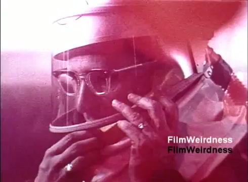 The still from 'Bell System Film from the 1970s' features a close-up of a person wearing a protective helmet and glasses, engaged in using a telephone. The color tone appears warm, possibly with pink or reddish hues, suggesting a vintage aesthetic. The individual is focused, emphasizing the functionality and innovation of telephone technology during that era. This moment captures a blend of industrial and personal communication history, evoking nostalgia for the time.