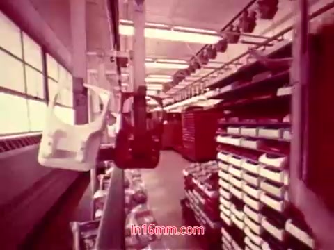 The still from the 16mm film 'Performance' (1971) shows an industrial setting, likely depicting a production line or workshop. The scene is rich in color, featuring shelving filled with various items, including partially assembled products. Elements like hangers and equipment suggest an organized workspace. The focus on machinery and materials aligns with the film's theme, emphasizing the intersection of technology and human activity. The end titles indicated give information about the production date and credits for the cast and crew involved.