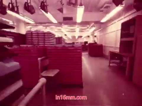 The still from the 16mm film 'Performance' - Bell System Film (1971) depicts a brightly lit interior space, likely a warehouse or storage area filled with stacked materials, possibly stacks of paper or equipment. The scene has a vivid pinkish hue, characteristic of the film's style, enhancing its visual uniqueness. This frame captures the end titles, which include the production date and names of the cast and crew, marking the film's conclusion.