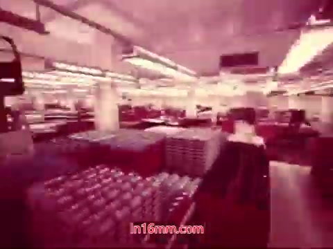 The still from the 16mm film 'Performance' (1971) features a vibrant, industrial setting. It captures a wide interior shot of a production area filled with stacks of containers or equipment. The lighting is bright, creating a pulsating ambiance, while the organized layout suggests a sense of efficiency. This image likely serves as a backdrop for the end titles, summarizing the film’s production details and credits.