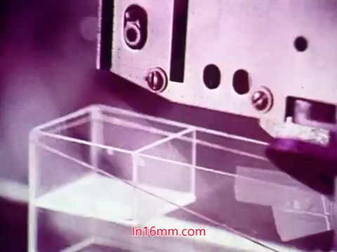 The still from the 16mm film 'Performance' (1971) captures a close-up of a mechanical device interacting with a clear plastic container. The machinery, possibly part of a production or assembly process, is highlighted with a purple tint, adding a stylized look. This scene likely serves as a visual representation of industrial or technological themes explored in the film. The end titles provide context, acknowledging the date of production and listing the cast and crew involved in the project.