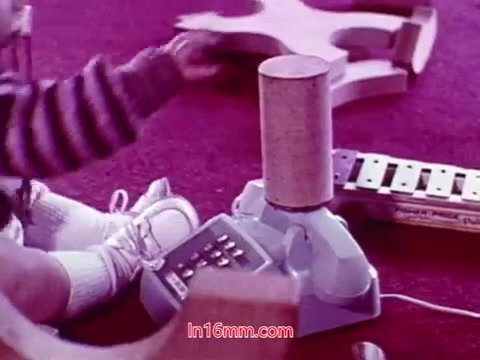 The still from the 16mm film 'Performance' presents a child engaging with a toy that resembles a small, colorful musical instrument. The child, dressed in a cozy outfit, sits on a vibrant carpet, surrounded by various play items. The focus is on the interaction between the child and the toy, capturing a moment of playfulness in a warm, nostalgic setting. The film, produced in 1971, features end titles that acknowledge the date of production and the cast and crew involved.