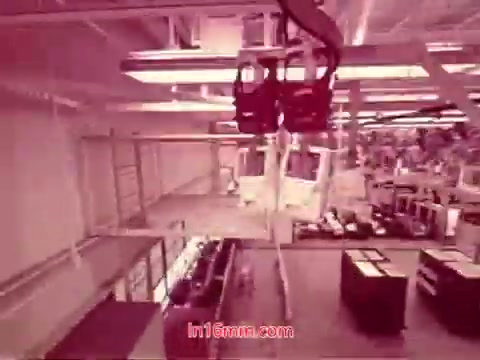 The 16mm still from 'Performance' is a scene showing a spacious industrial setting, possibly a factory or workshop. Overhead, there are mechanical devices or cranes positioned, suggesting an assembly or manufacturing process. The lighting is bright and artificial, contributing to a somewhat sterile atmosphere. The overall color is muted, leaning towards a pinkish hue. This still likely represents the set design and operational context of the film, focusing on the technological themes involved in production, with an emphasis on machinery and space.