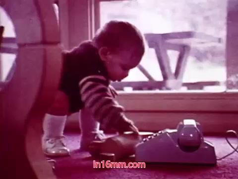 The still from 'Performance' shows a young child interacting with a vintage telephone, set against a backdrop of a sunlit room. The child is dressed in a striped sweater and white socks, displaying curiosity as they lean over the phone on a textured carpet. The composition highlights the innocence and exploration of childhood within a cozy, domestic setting.