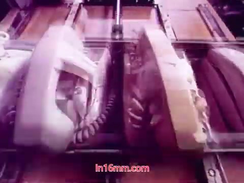 The still from 'Performance,' a Bell System Film from 1971, features a close-up of two vintage telephone models, one in white and the other in beige, arranged in a mechanical or production setting. The image captures the telephones in a factory-like environment, emphasizing their design and technological context. The vibrant colors and textures reflect the period's aesthetic, drawing attention to the product's significance in communication history. The end titles, likely displayed at the bottom, provide details about the production date and the cast and crew involved.