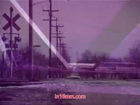 The still from the 16mm film 'Performance' - Bell System Film (1971) features a desolate landscape with utility poles lining a gravel road. A train crossing sign indicates nearby railway tracks. The scene is bathed in a purple hue, adding an abstract quality. It captures a moment of stillness, presumably part of the film's concluding credits, conveying a sense of nostalgia and atmosphere.