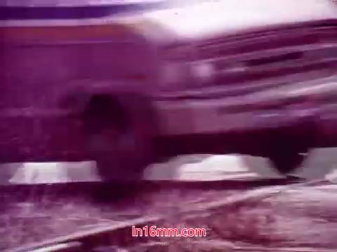 The still from 'Performance' - Bell System Film (1971) features a close-up of a vehicle, likely a van, moving rapidly along a track. The image is characterized by a distinct color palette, with a pinkish or purplish hue dominating the scene. This gives it a somewhat surreal or altered appearance, typical of the film's stylistic choices. The movement conveys a sense of urgency, fitting the film's themes. The end titles, which include production details and the cast and crew, are likely displayed across the frame, summarizing the film's credits.