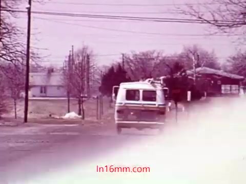 The 16mm still from 'Performance' - Bell System Film (1971) captures a scene featuring a white van driving down a rural road. The background features sparse trees and houses, indicative of a quiet setting. The color palette showcases muted tones, typical of the era, with a slight haze suggesting a nostalgic or dreamlike atmosphere. This image likely serves as a transitional moment in the film, bridging the narrative with the end titles that include production details and credits.