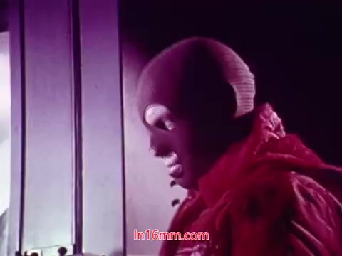 The still from the film 'Performance' features a character wearing a mask and a vibrant red outfit. The background appears to have a mix of colors, likely contributing to the film's stylistic approach. The image captures a moment that seems to convey a sense of intrigue or drama. As this is a frame from the end titles, it includes details such as the production date and credits for the cast and crew involved in the film.