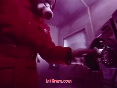 The still from 'Performance' showcases a figure in a red suit and gas mask, emphasizing a surreal and dystopian atmosphere. The environment appears industrial, with a telephone prominently featured. The use of purple hues adds a sense of unease, typical of the film's avant-garde style. This moment likely serves as a poignant conclusion to the narrative, aligning with the themes of communication and isolation present in the film.