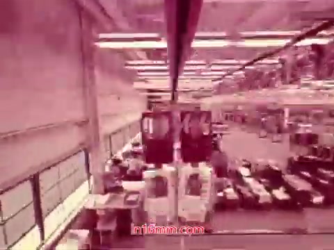 The still from the 16mm film 'Performance' - Bell System Film (1971) likely depicts an industrial or manufacturing setting, showcasing a spacious interior with visible machinery and assembly lines. The lighting creates a distinct atmosphere, possibly with a pinkish hue, typical of older film prints. The end titles provide essential production details, including the date and the cast and crew involved. This scene marks a significant moment, concluding the film with a sense of completion and context.