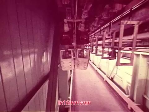 The still from the 16mm film 'Performance' features an industrial setting, characterized by an overhead view of a production area. The focus is on suspended structures, likely part of a manufacturing or assembly line, with visible machinery and equipment. The color palette leans toward a reddish tint, adding a unique aesthetic. This moment captures the conclusion of the film, showcasing end titles that provide production details, cast, and crew information, highlighting its 1971 origins.