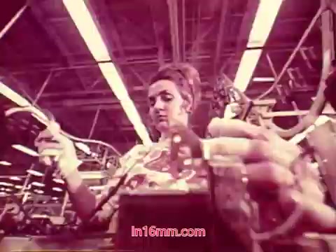 The still from the Bell System film 'Performance' (1971) depicts a woman working in a factory setting. She is focused on her task, holding tools or components, amidst a backdrop of machinery and assembly lines. The image is likely in muted colors, typical of 16mm prints from that era. This moment captures the industrious environment of the time, highlighting both the labor process and the individuals involved. The film's end titles convey production details and recognize the cast and crew, marking the conclusion of the production.